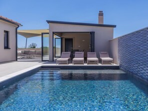 A modern outdoor pool area with clear blue water, a covered seating area, and a small building with glass doors.