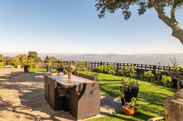 An outdoor patio with a wicker table and chairs, surrounded by a well-maintained garden and a view of the water.