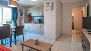 A modern kitchen with white cabinets, a microwave, and a coffee maker. There is a dining area with a table and chairs, and a TV on a wooden stand. - Les Thermes 532 - Vue mer avec piscine partagée (Concarneau)