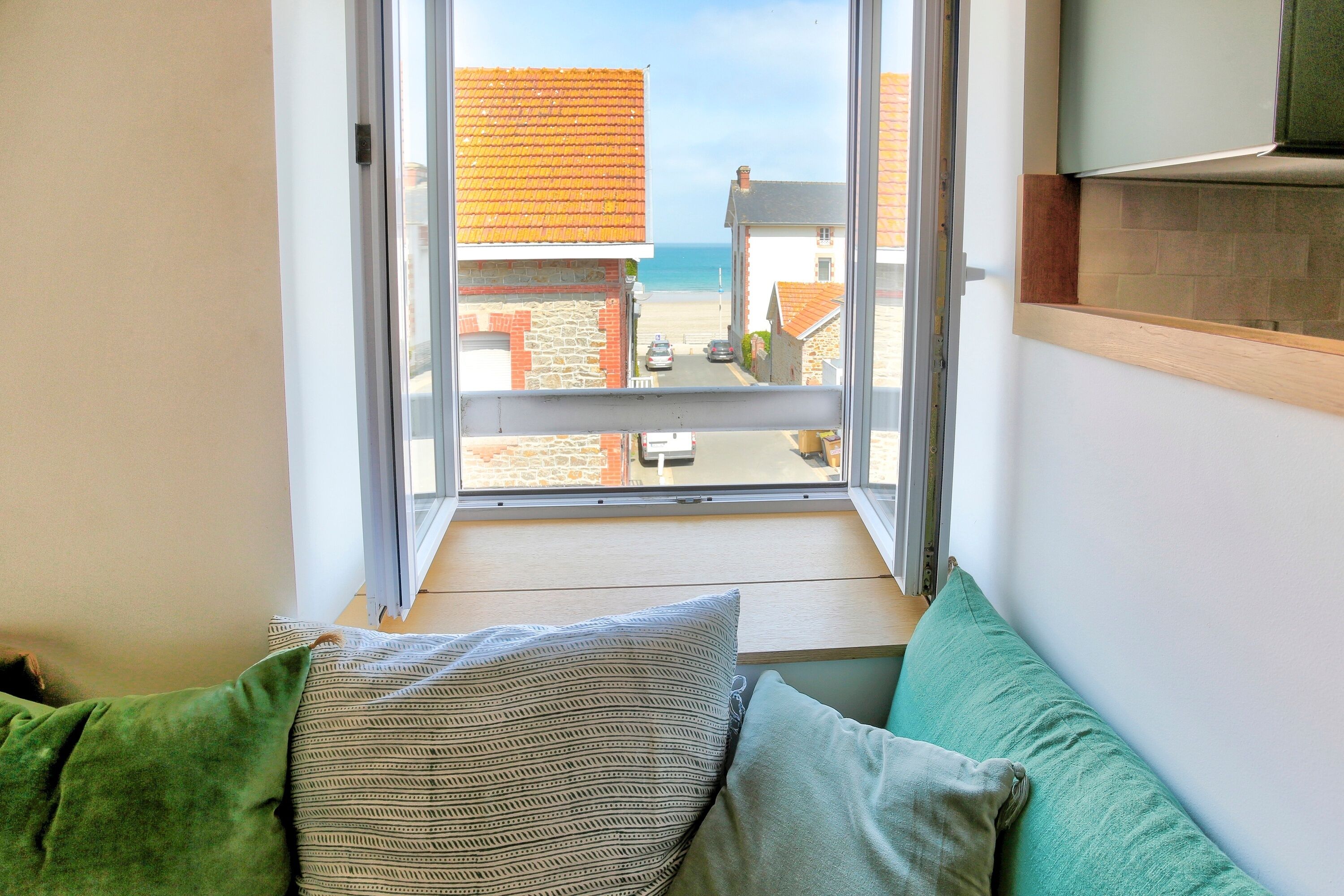 Ein Zimmer mit Fensterblick auf eine Straße und Gebäude, ein grünes Sofa mit gemusterten Kissen und ein Holzregal.