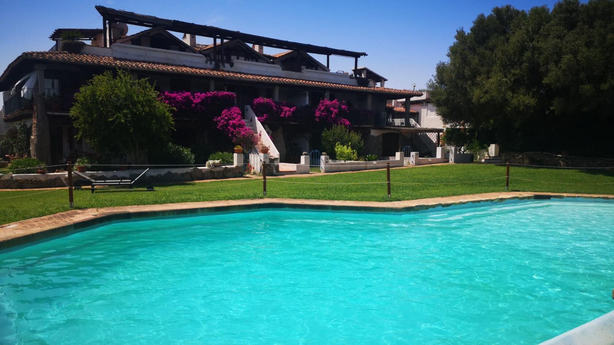 Une piscine à l’eau d’un bleu limpide, entourée d’une pelouse bien entretenue et d’un bâtiment doté d’un toit en tuiles et de balcons.