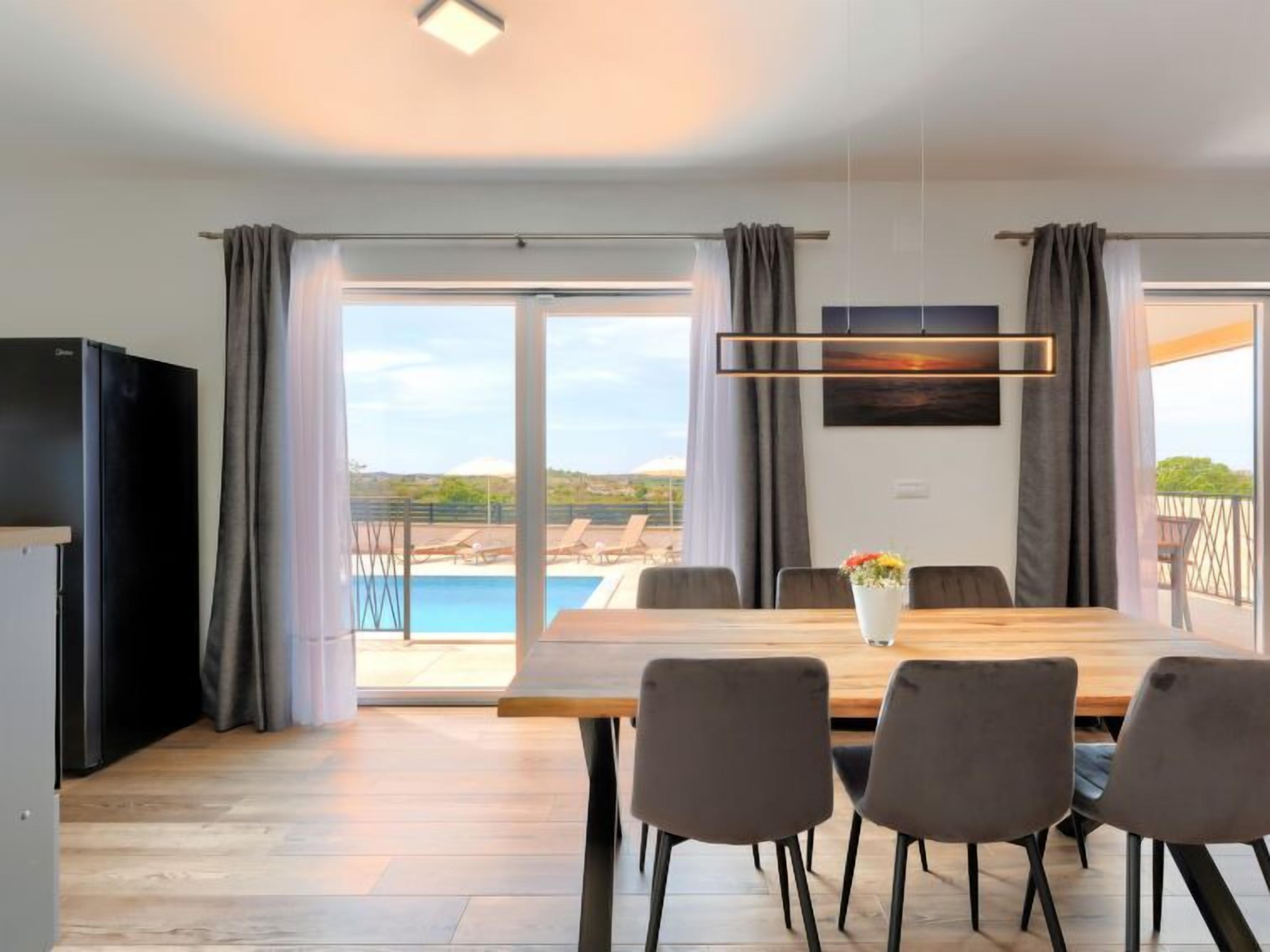 Un comedor moderno con una mesa de madera, cuatro sillas y vistas a una piscina y paisaje a través de amplios ventanales.