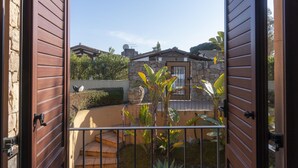 Vista a través de puertas de madera abiertas que dan a un patio con un muro de piedra, un pequeño jardín con plantas y un edificio con techo de tejas al fondo.