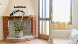 Quarto com lareira de tijolos, um vaso de planta, sofá branco e uma porta com persianas verticais.