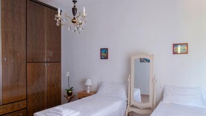Quarto de hotel com duas camas, um guarda-roupa de madeira, um candelabro, uma mesinha de cabeceira com uma lâmpada e duas fotografias emolduradas na parede.