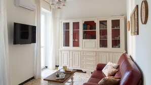 Un salon comprenant un téléviseur à écran plat, une table basse avec des livres et une tasse, un canapé rouge orné de coussins à motifs et un meuble blanc aux portes en verre.