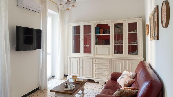Un salon comprenant un téléviseur à écran plat, une table basse avec des livres et une tasse, un canapé rouge orné de coussins à motifs et un meuble blanc aux portes en verre.