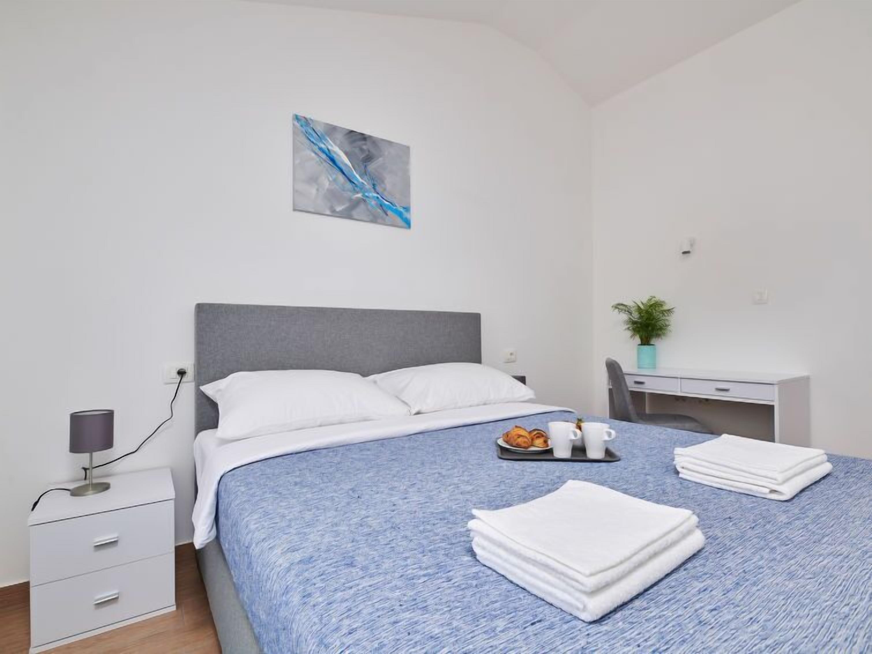 A neatly made bed with white linens, a gray headboard, and a blue textured bedspread. There is a small bedside table with a lamp, a white drawer, and a wall-mounted power outlet. A desk with a chair and a potted plant is visible in the corner.
