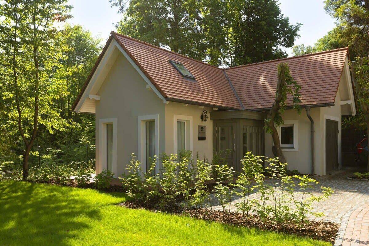 Une maison de plain-pied avec un toit en pente, un porche à l’avant et un petit jardin planté de jeunes arbres.