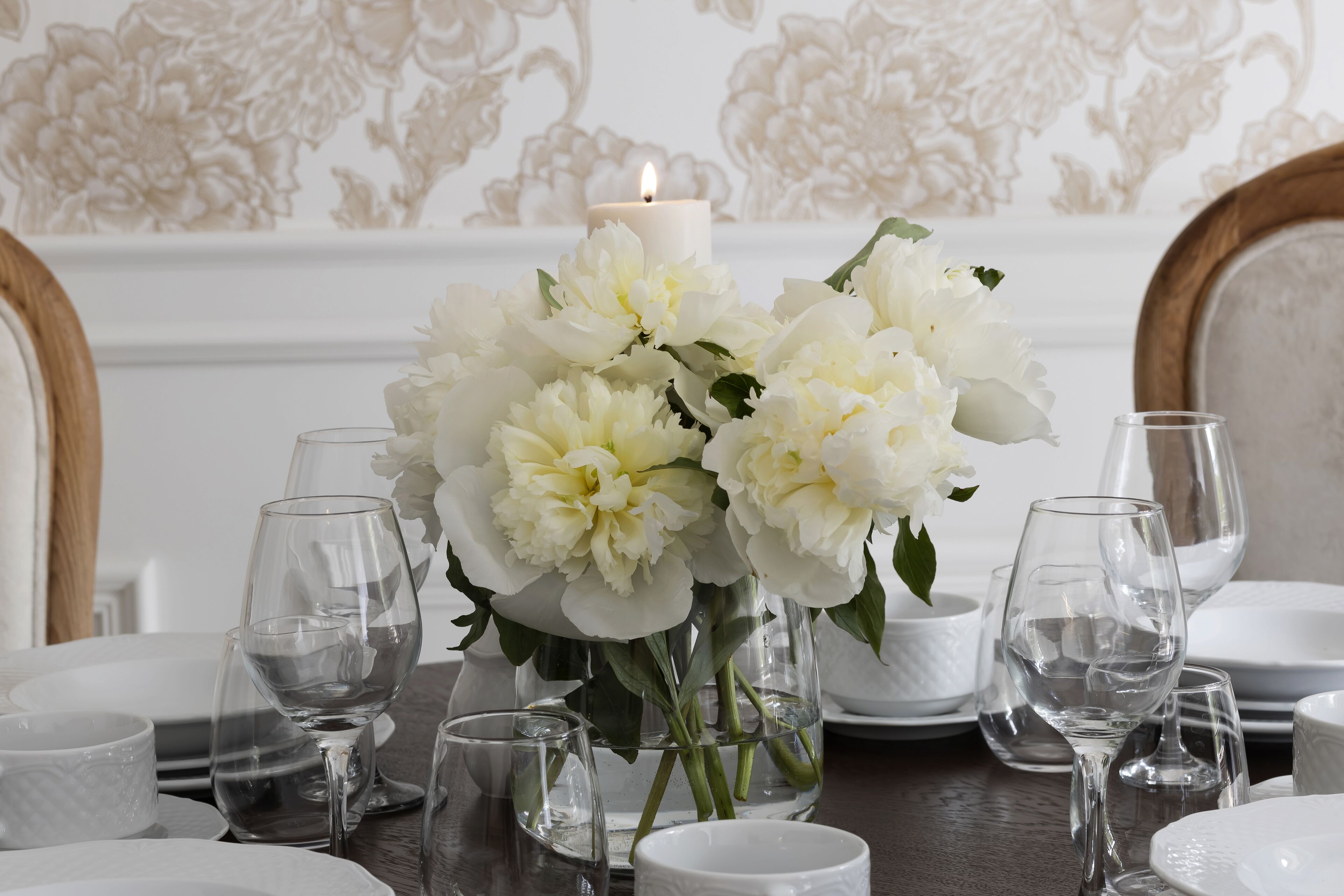 A dining table set with white plates, clear wine glasses, and a vase with white flowers.