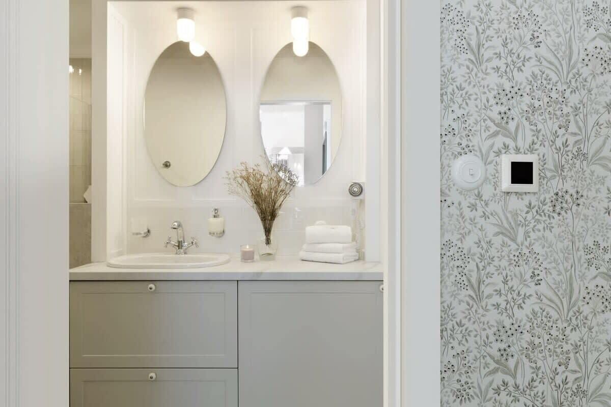 A bathroom with a double sink, two mirrors, a vase with dried flowers, and a towel rack.