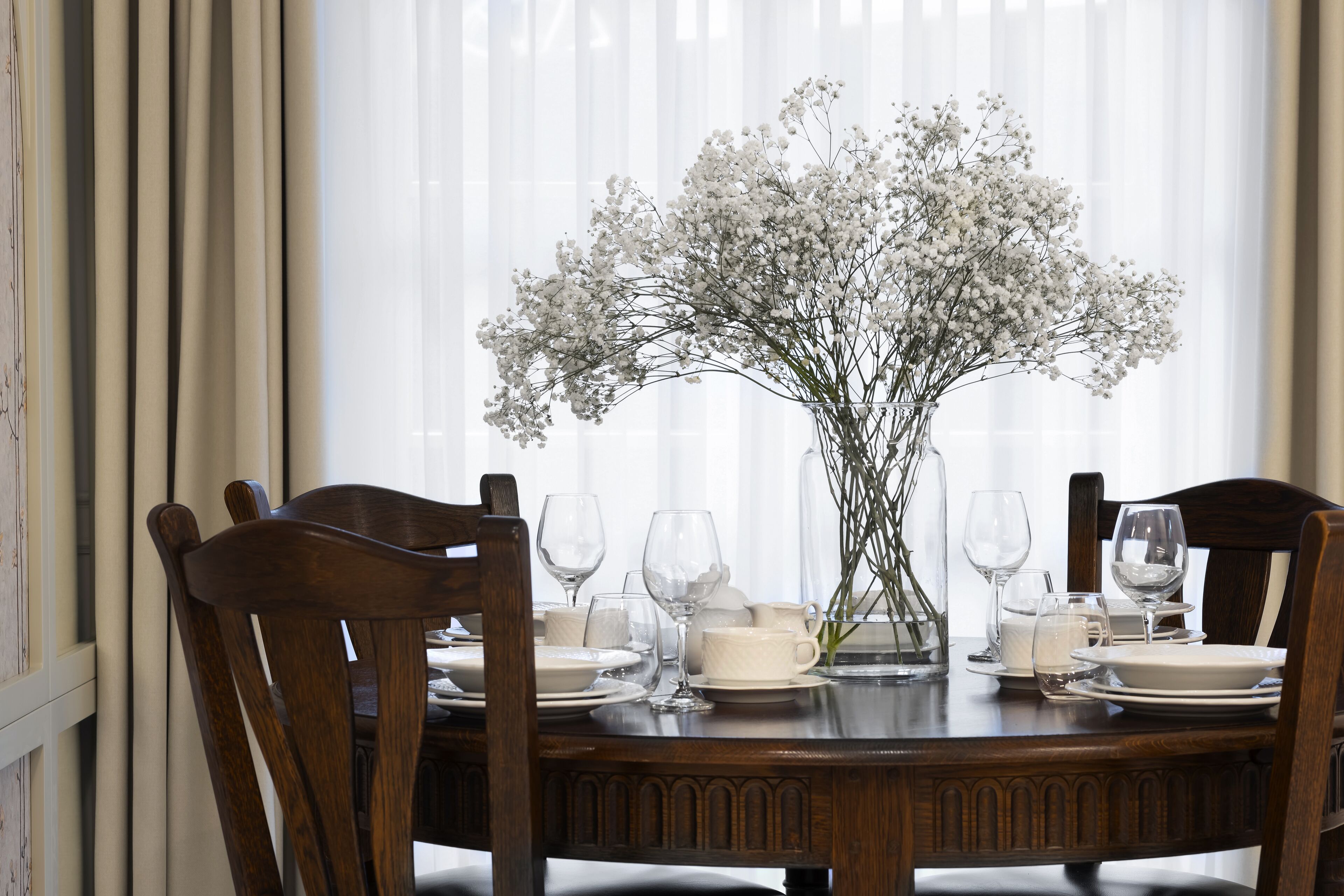 Een eettafel gedekt met witte borden, glazen en een vaas witte bloemen.