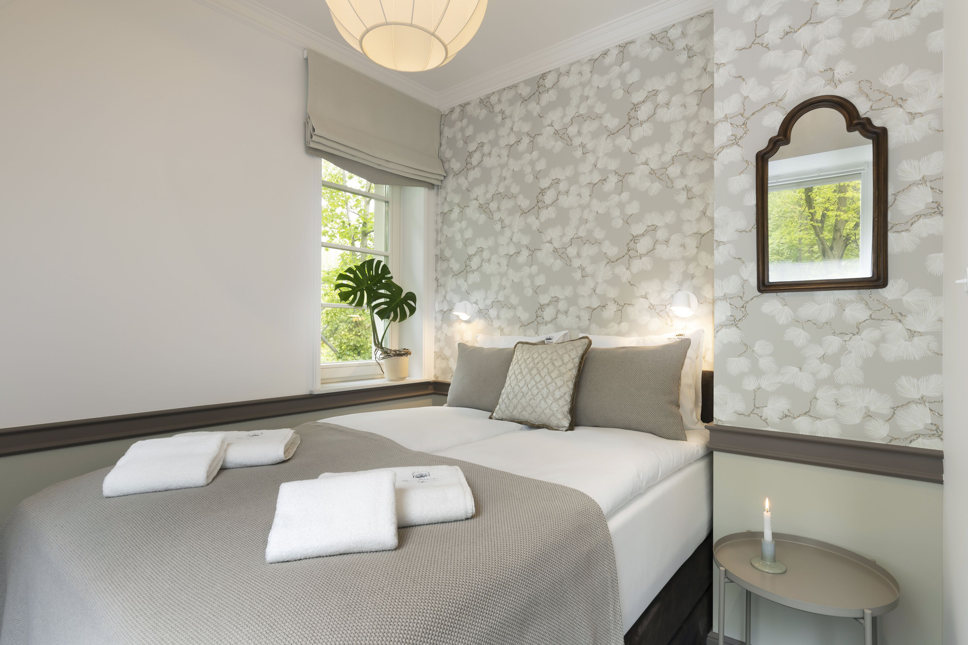 A neatly made bed with towels, a bedside table with a candle, and a window with a plant.