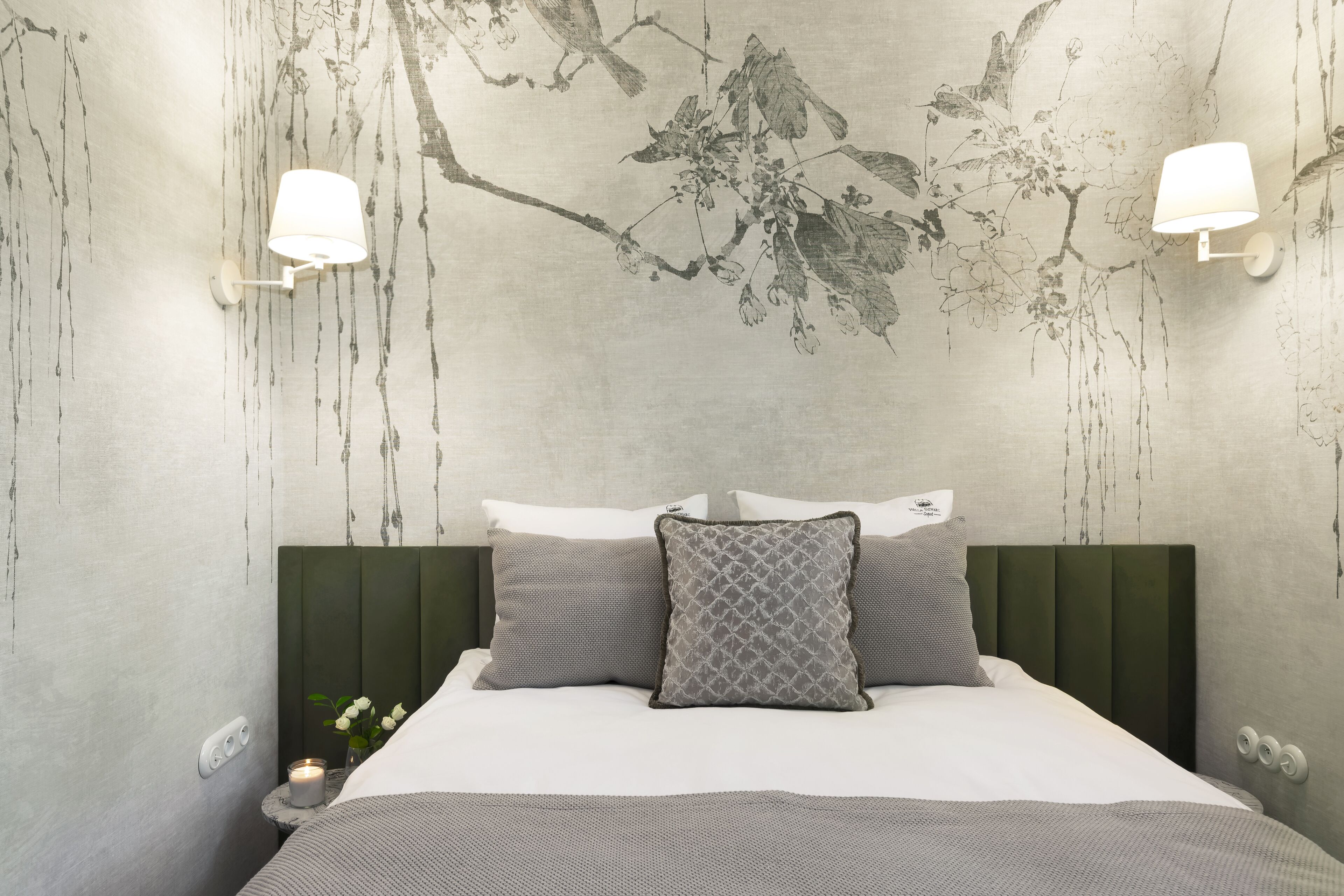 A neatly made bed with a green headboard, two wall-mounted lamps, and a wall with a floral pattern.