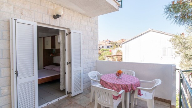 Un balcone con tavolo e sedie, una porta socchiusa che dà su una stanza, e un edificio bianco sullo sfondo.