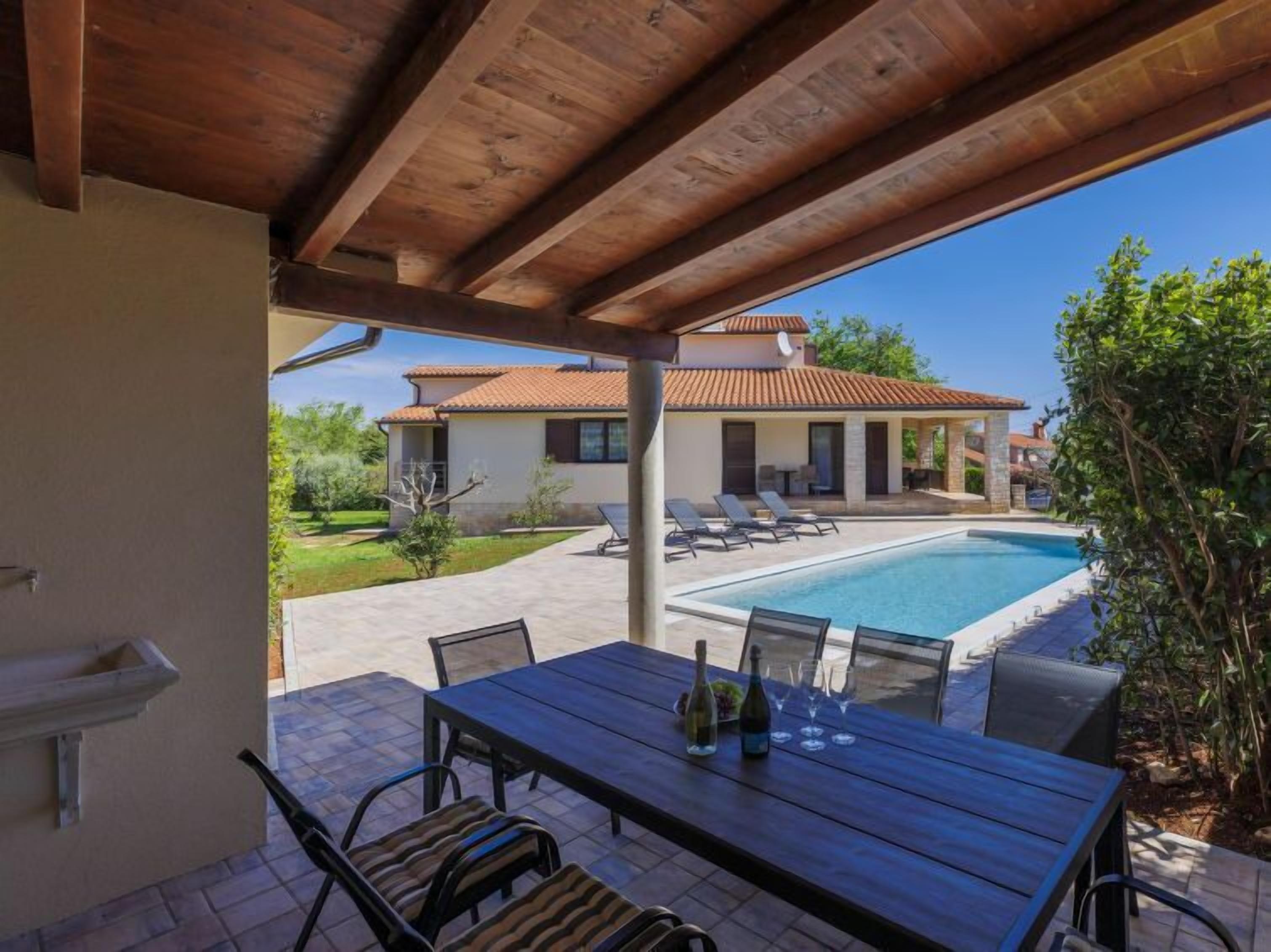 A covered patio with a pool, outdoor seating, and a view of a house with a tiled roof.