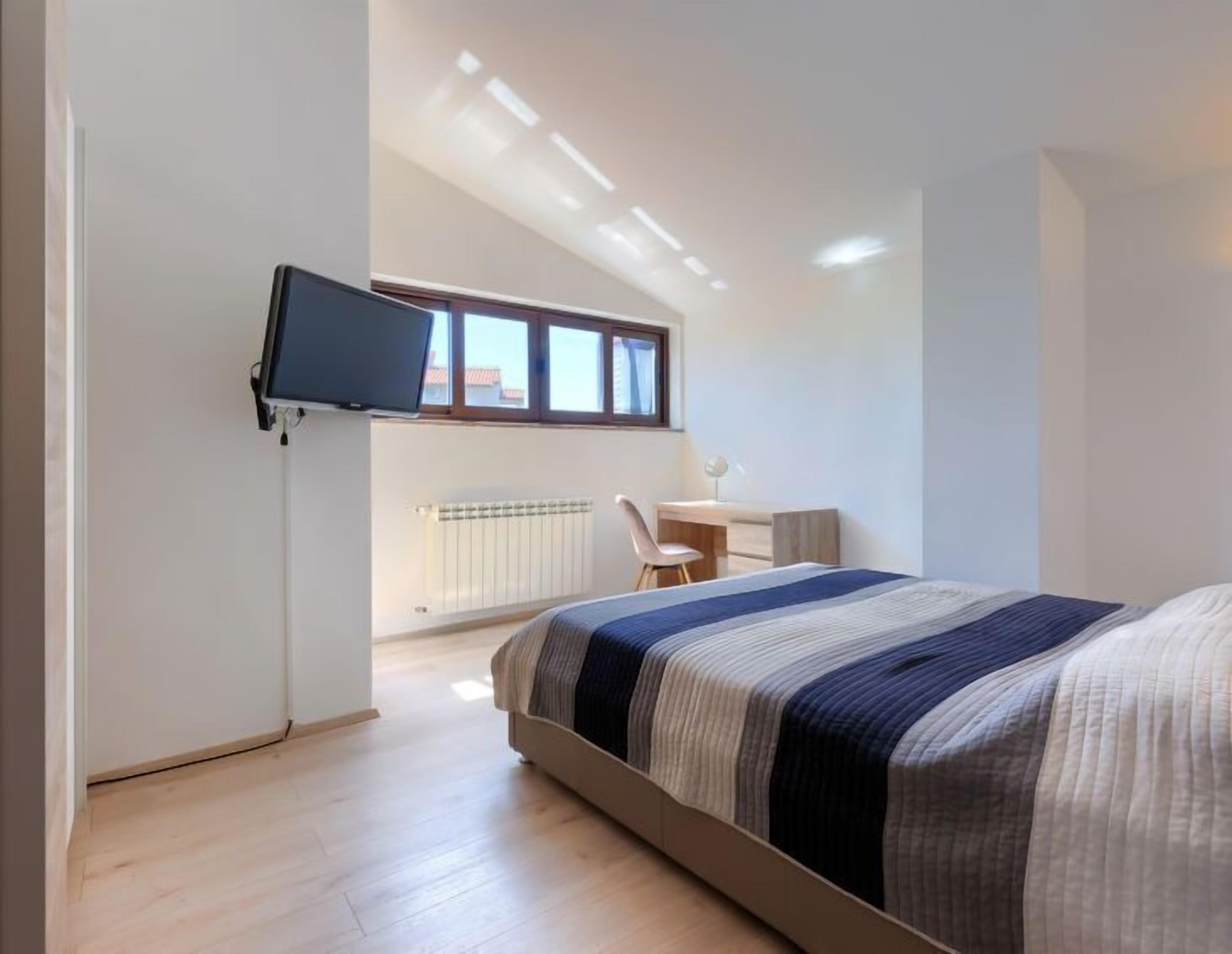 Ein Schlafzimmer mit einem Bett, einem Schreibtisch, einem Sessel, einem an der Wand montierten Fernseher und einem Fenster.