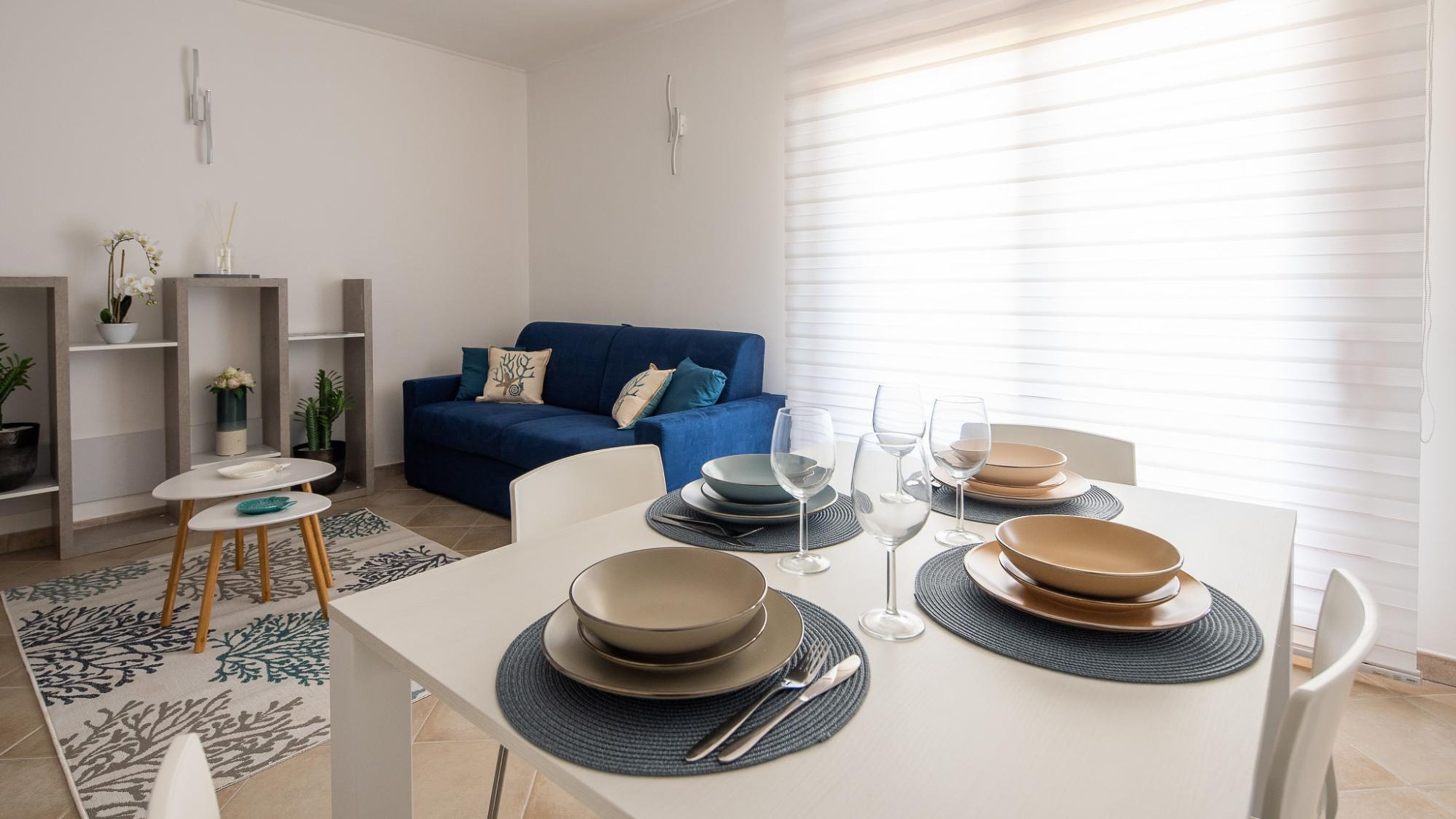 A modern living room with a dining area. There is a blue sofa with cushions, a small side table with a vase and a plant, and a dining table set with plates, bowls, and glasses.