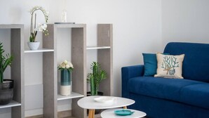A modern living room with a blue sofa, white coffee table, and shelves with plants and decorative items. - Welcomely - Xenia - Attico Supramonte (Olbia)
