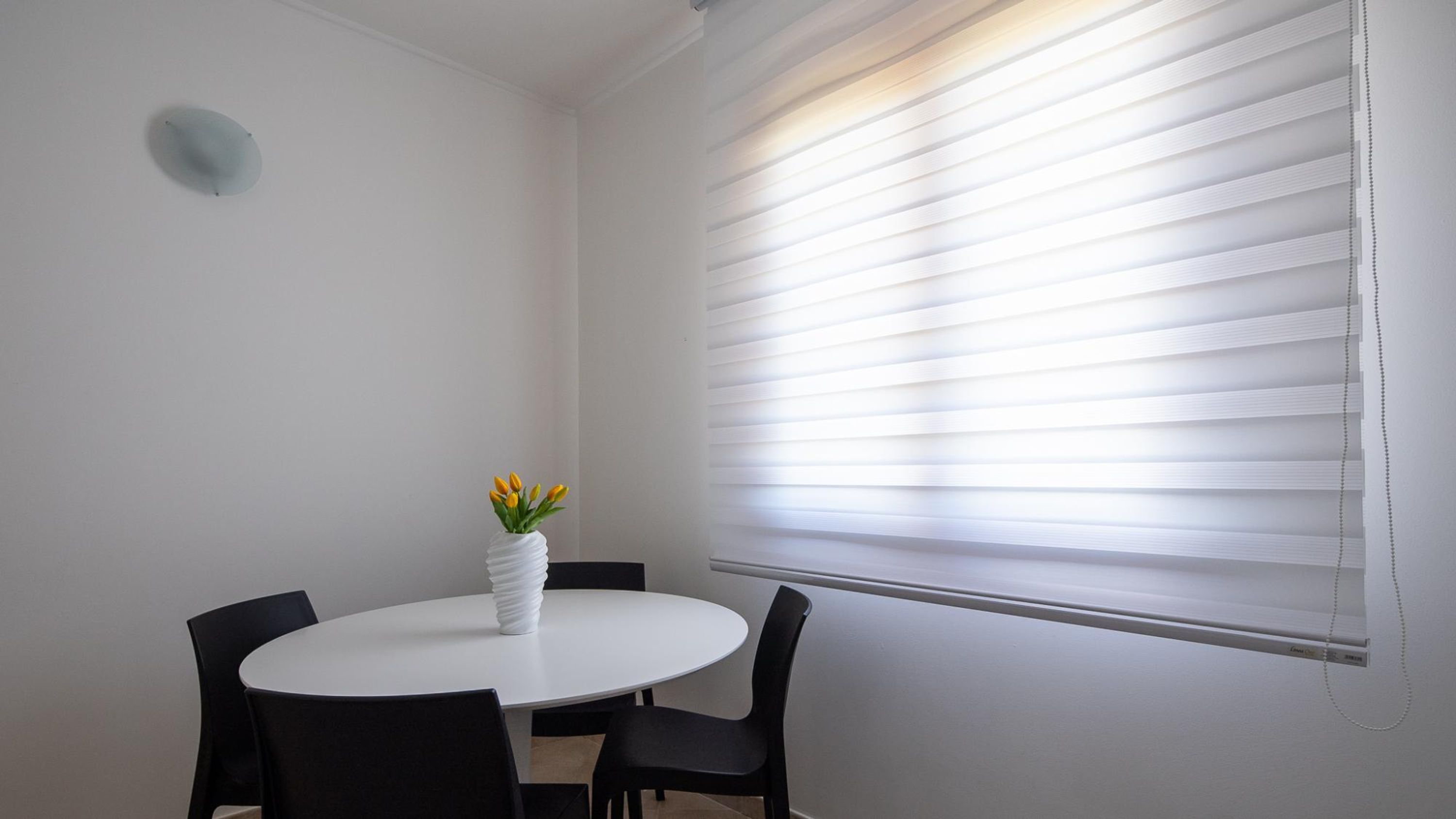 A dining area with a round table, four chairs, a white vase with yellow flowers, and a window with blinds.