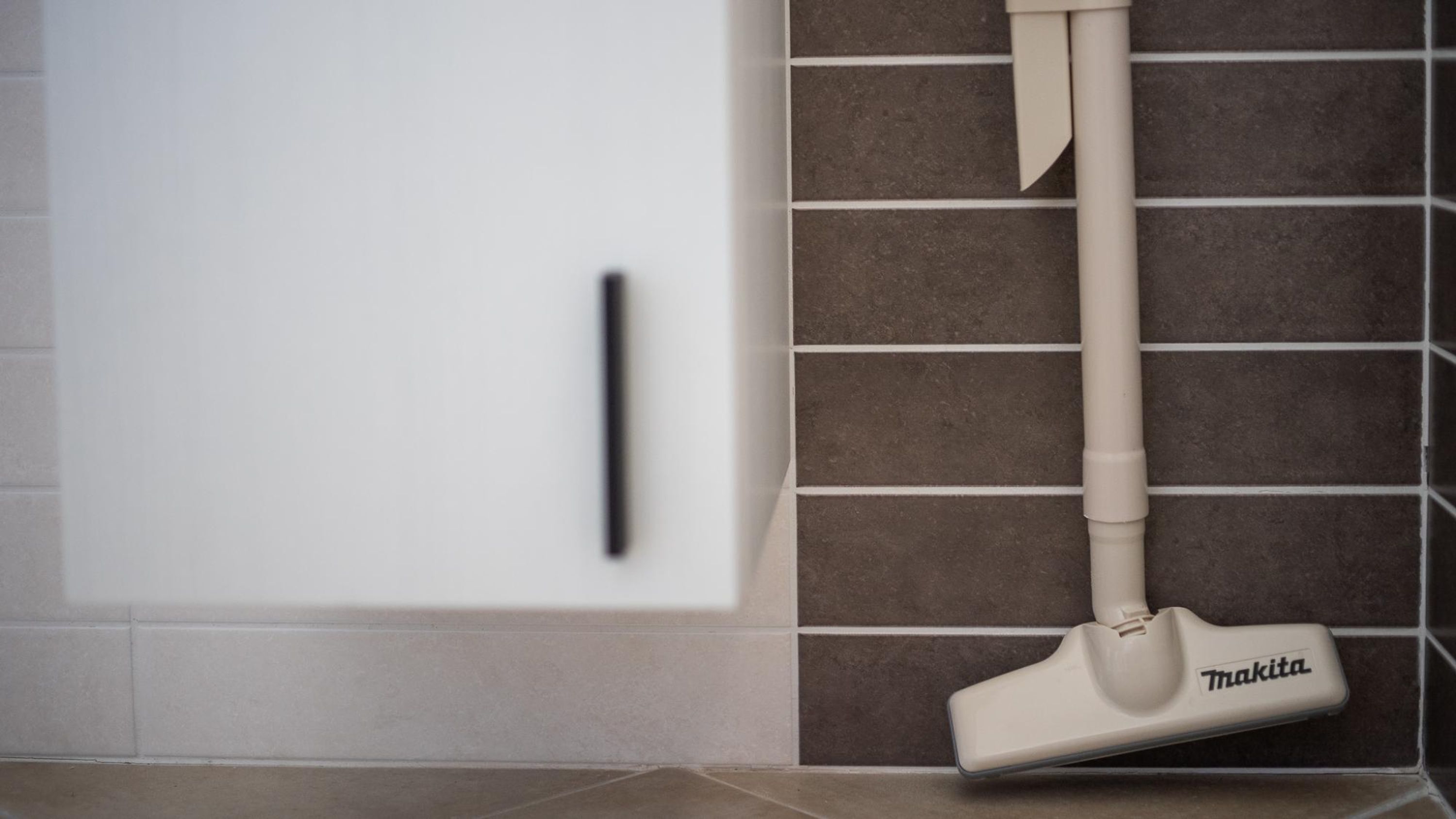 A vacuum cleaner with a Makita brand label, positioned against a tiled wall with a white cabinet and a black handle.