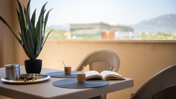 A balcony with a table set for a meal, a potted plant, a book, and a thermos. - Welcomely - Xenia - Attico Supramonte (Olbia)