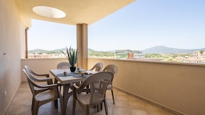 A balcony with a table and chairs, a potted plant, and a view of buildings and mountains. - Welcomely - Xenia - Attico Supramonte (Olbia)