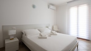 A minimalist hotel room with a large bed, white linens, a bedside table with a lamp, and a window with blinds. - Welcomely - Xenia - Attico Supramonte (Olbia)