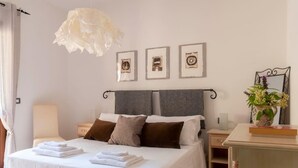 Een net opgemaakt bed met witte lakens en gevouwen handdoeken, een nachtkastje met een lamp, en een muur met drie ingekaderde foto's.