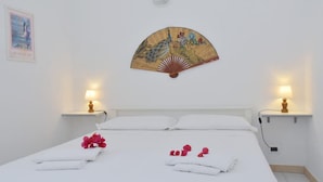 A neatly made bed with white linens and pink flowers, two bedside lamps, and a decorative fan on the wall. - Welcomely - Villa Oleandro (Muravera)