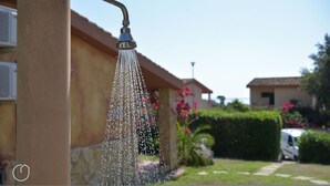 A showerhead with water flowing, a house with a stone facade, and a vehicle parked in the background. - Welcomely - Villa Oleandro (Muravera)