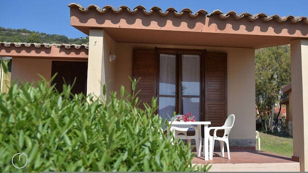 A building with a tiled roof, a covered patio area with a table and chairs, and a view of greenery and hills in the background. - Welcomely - Villa Oleandro (Muravera)