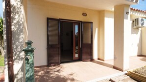 Entrada de un edificio con puerta y contraventanas de madera, un pequeño espacio exterior con piso de baldosas y un equipo de aire acondicionado en la pared.
