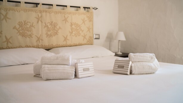 Una cama bien hecha con sábanas blancas y dos toallas, una mesita de noche con lámpara, y un cabecero decorativo.