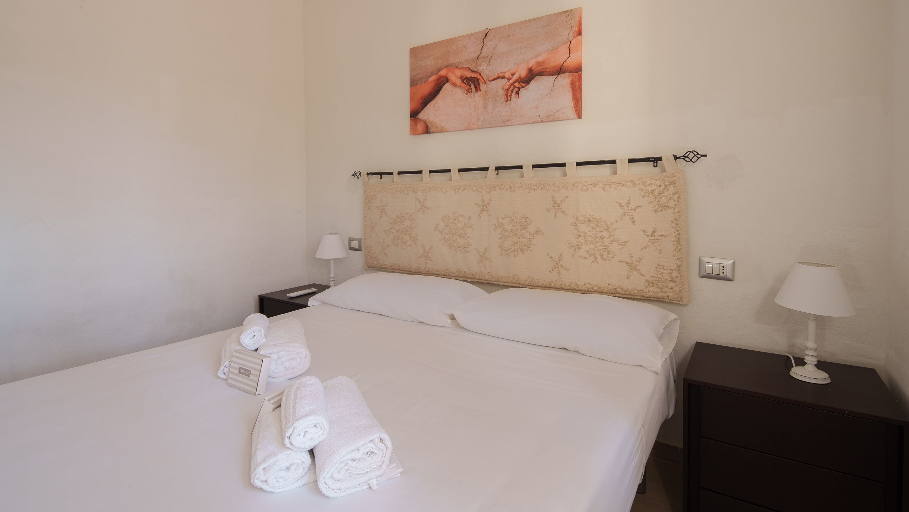 A neatly made bed with white linens, a headboard with a floral pattern, and a painting above it.