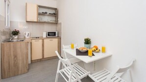 Een compacte keuken met een magnetron, spoelbak en houten kastjes. Er is een eettafel met stoelen en een kleine potplant.