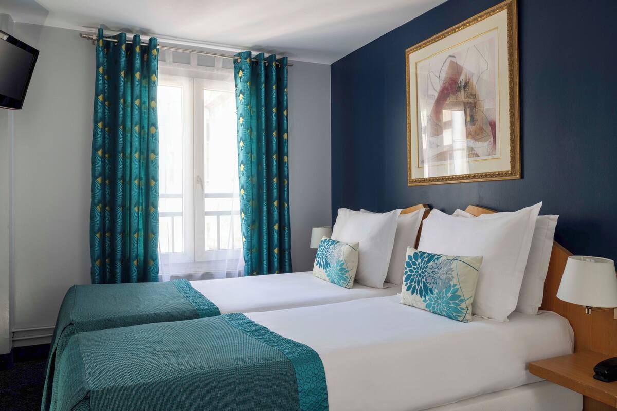 A hotel room with two beds, teal curtains, a framed artwork on the wall, and a bedside table with a lamp.