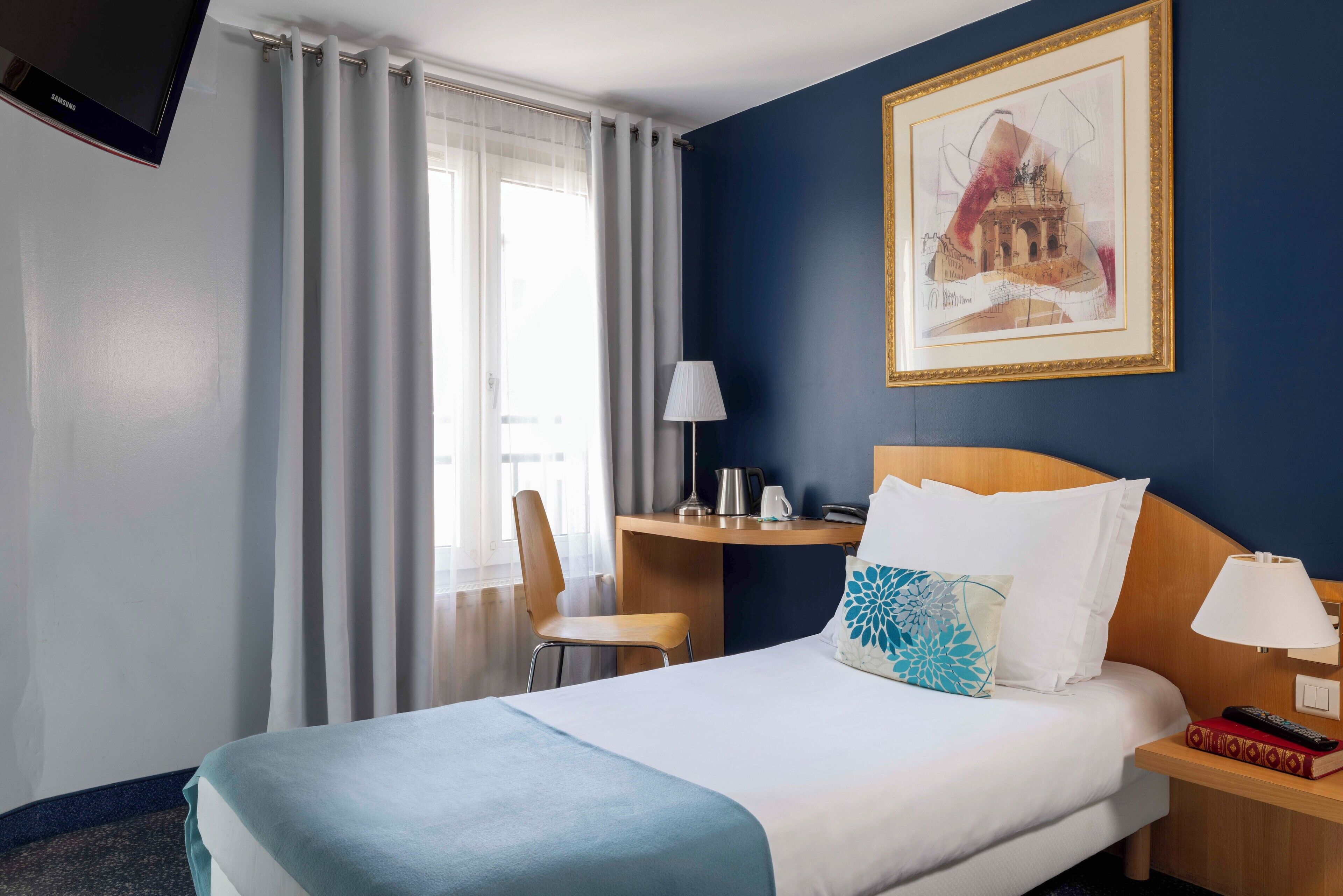 A hotel room with a bed, a desk, a chair, a TV, and a painting on the wall.