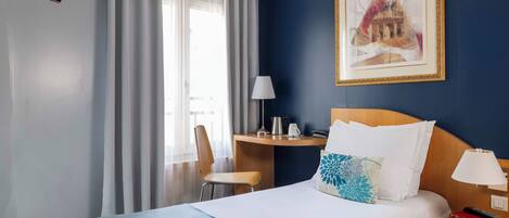A hotel room with a bed, a desk, a chair, a TV, and a painting on the wall.