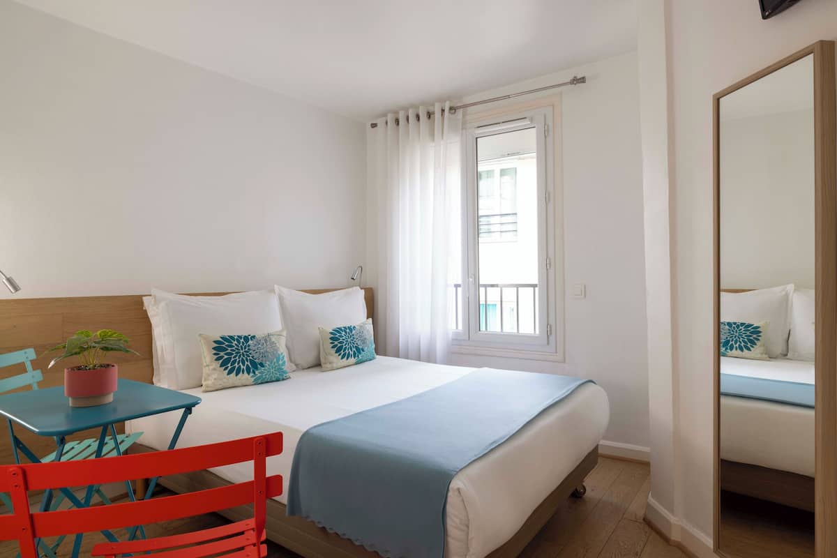 A bedroom with a bed, a small table with a plant, a chair, and a mirror.