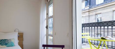 Una habitación con una cama, una silla morada, una mesita con un periódico y vistas a un edificio a través de una ventana.