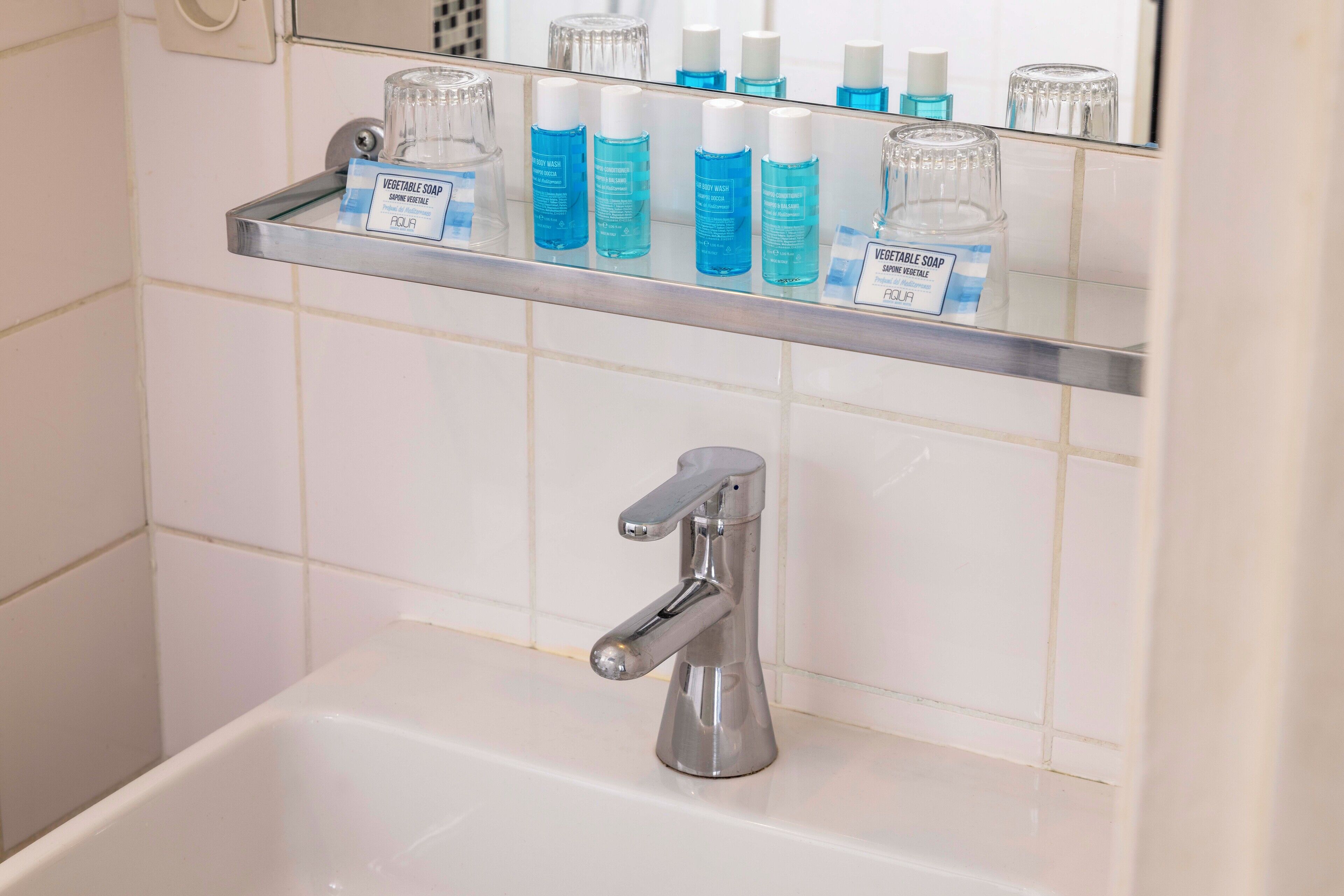 A bathroom with a chrome faucet, a shelf with blue bottles of vegetable soap, and a mirror above the sink.
