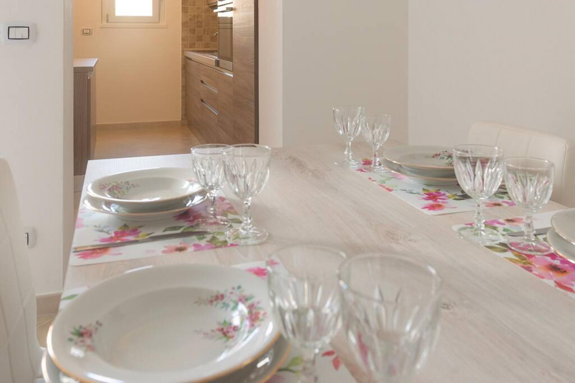 Une table à manger dressée avec des assiettes et des verres, un chemin de table à motifs floraux, et un coin cuisine en arrière-plan.