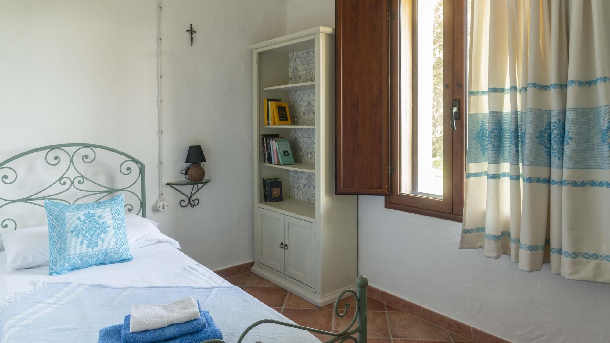 A bedroom with a bed, a bedside table with a lamp, a bookshelf, and a window with curtains.