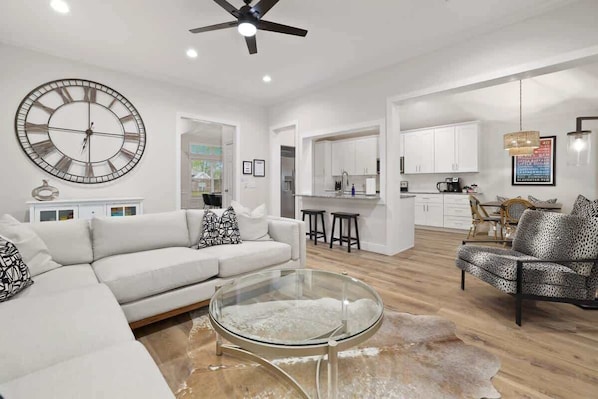 A modern living room with a glass coffee table, a large wall clock, and a ceiling fan.