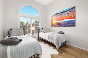 A bedroom with two beds, a large window, and a painting on the wall.