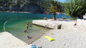 A pebbled beach with a clear blue sea, a palm tree, and a rocky hillside in the background. - Villa Riva - Five Bedroom Villa With Pool and Sea View (Dubrovnik)