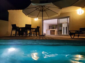 Área de piscina ao ar livre com deck de madeira, mesa com cadeiras e um guarda-sol.