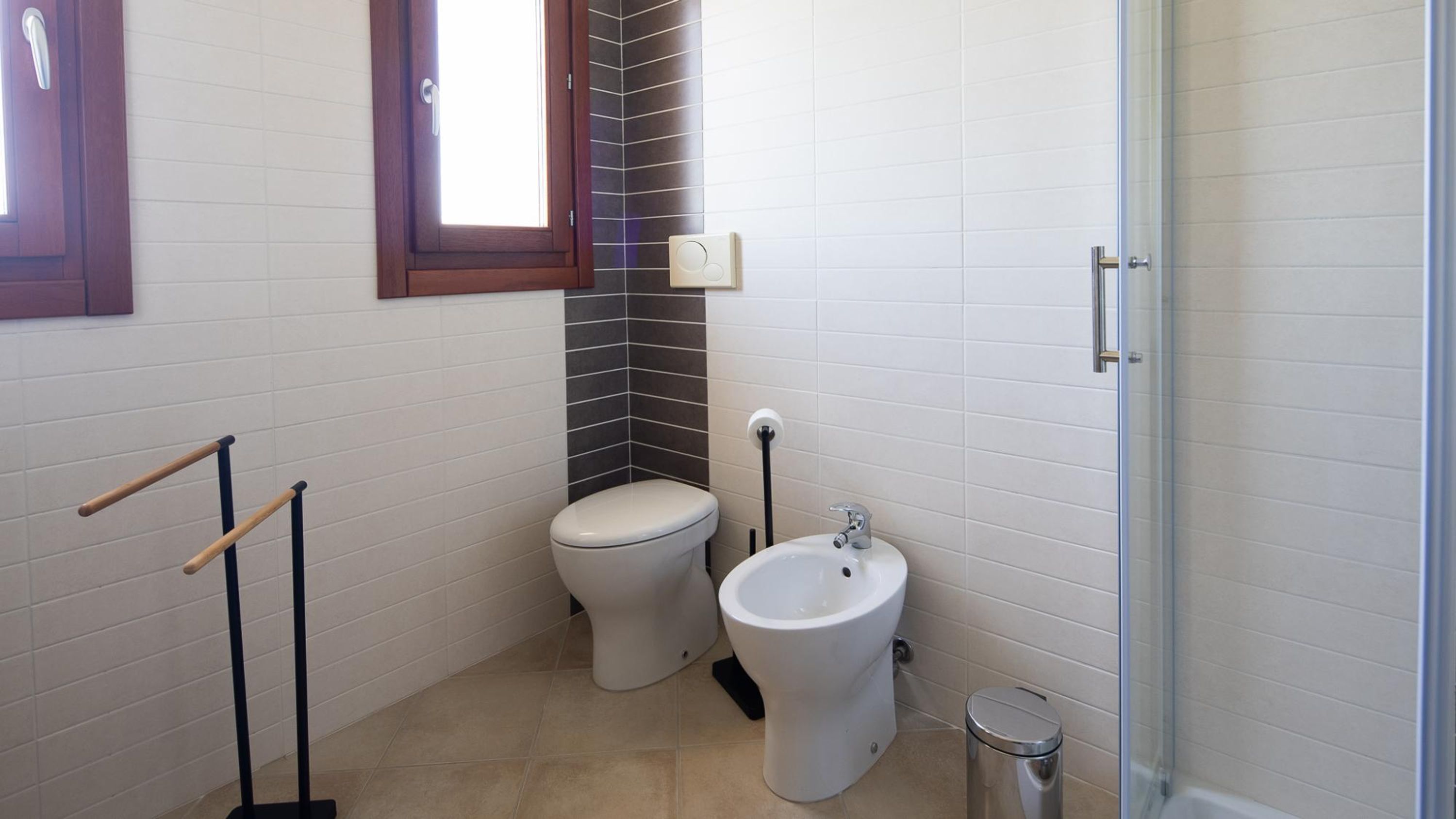 A bathroom with two toilets, a bidet, a trash bin, and a window with a wooden frame.