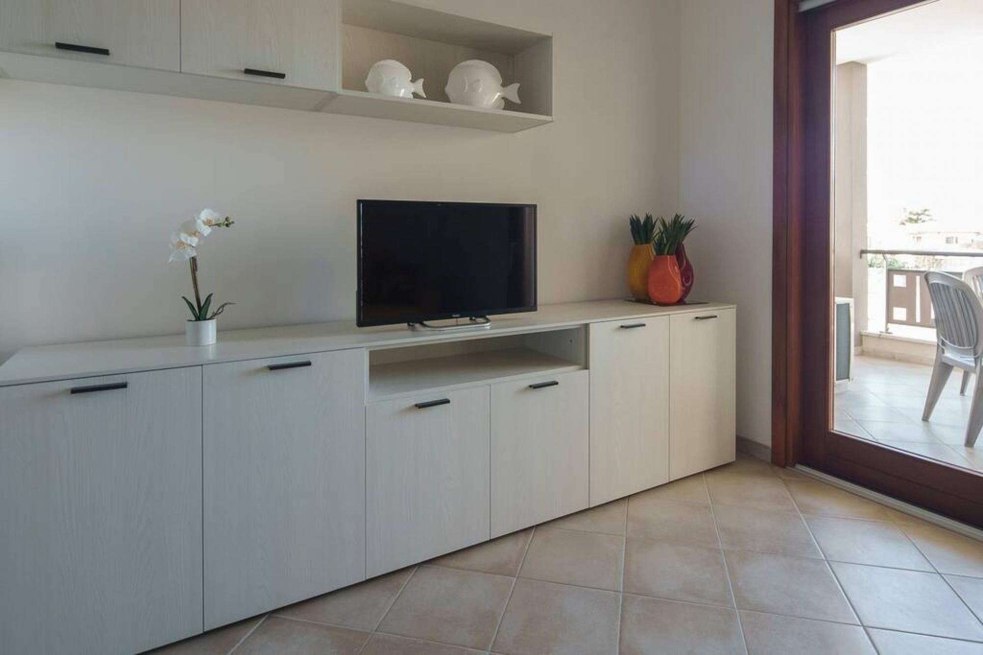 Una sala moderna con un televisor de pantalla plana sobre un mueble blanco, una pequeña orquídea en maceta y esculturas de peces decorativas en una estantería.